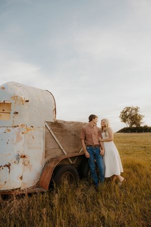 A couple stands together by a rustic vintage trailer in a golden field, sharing a tender moment outdoors.