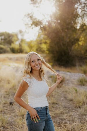 Smiling blonde woman in white lace top and jeans poses outdoors in golden sunlight with trees and dry grass.