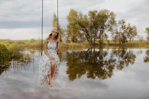 Woman in floral dress sitting on a rope swing over a reflective lake surrounded by lush green trees.