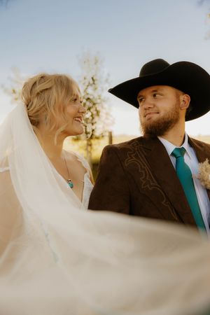 A bride in a flowing veil smiles at a bearded groom wearing a black cowboy hat and teal tie outdoors.