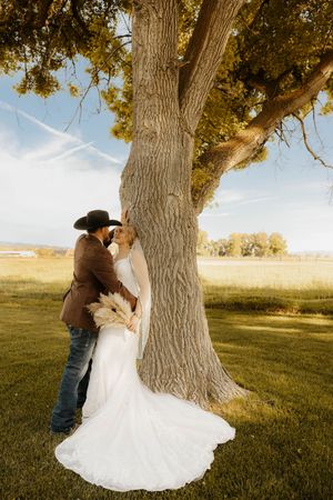 A bride and groom share a romantic kiss beside a large tree in a sunlit field, with the bride holding a boho bouquet.