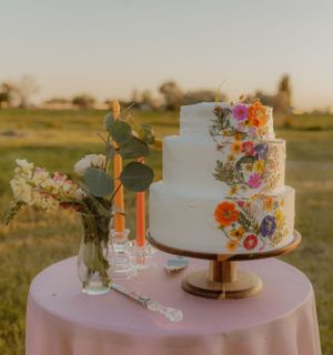 Wedding cakes decorated with fresh flowers displayed on pink tablecloth outdoors.