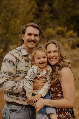 A happy family of three poses outdoors in fall, father with mustache holding smiling toddler beside blonde mother.