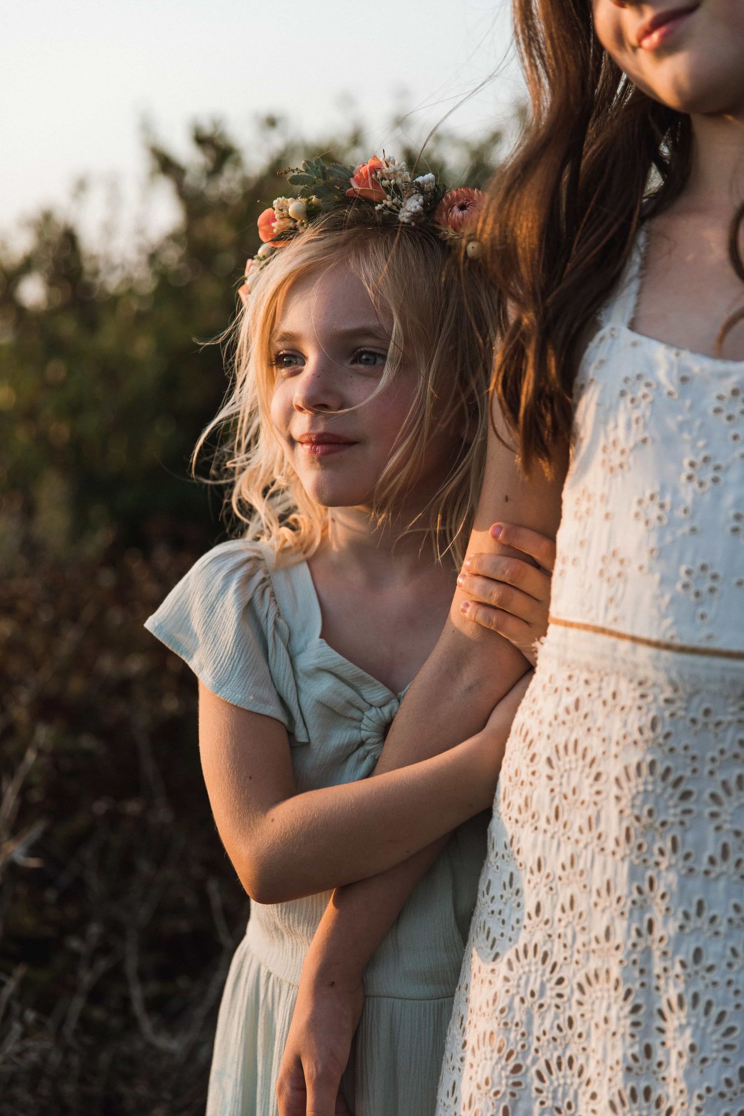 Young blonde girl wearing floral crown hugging mother in white lace dress at golden hour outdoors.