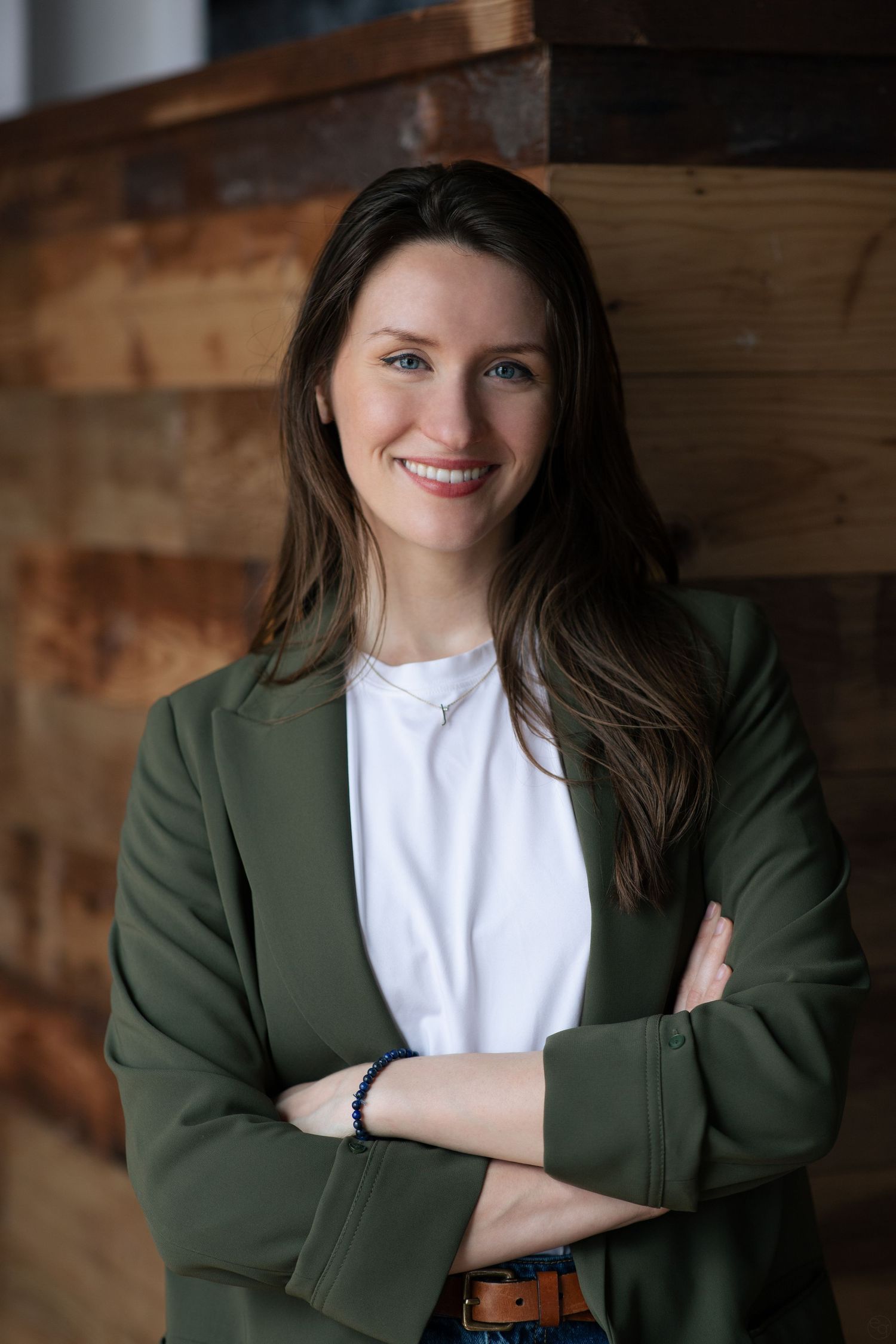 Professional portrait of someone in a green blazer and white t-shirt standing against a wooden wall background.