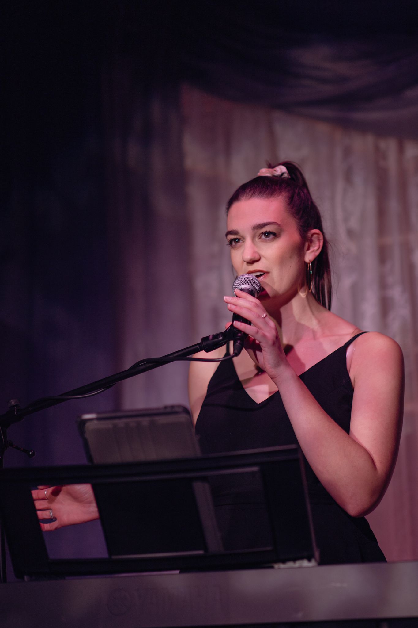 A performer in a black tank top singing into a microphone while standing at a keyboard on a dimly lit stage.