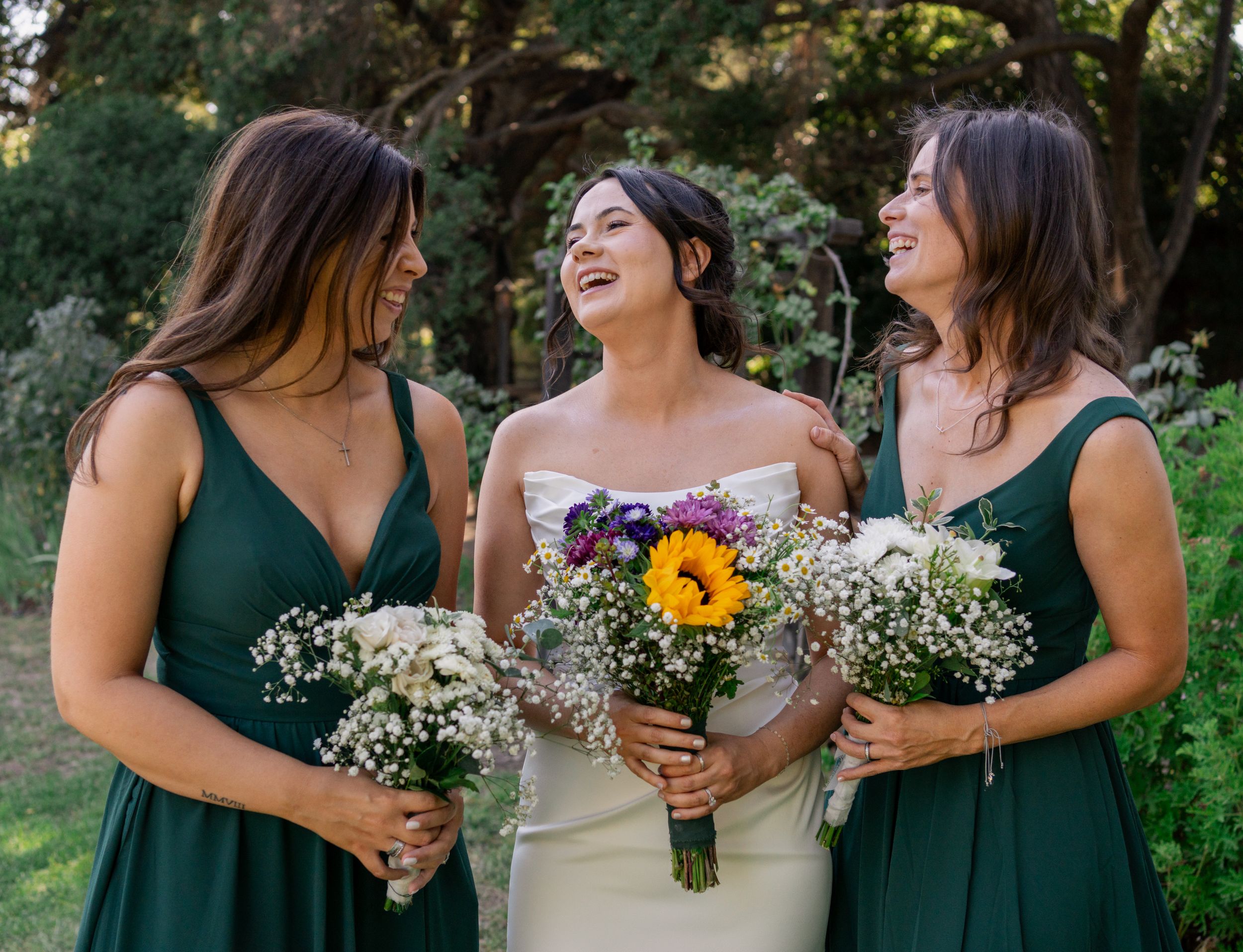 Bride in ivory gown laughs with two bridesmaids in green dresses holding wildflower bouquets.