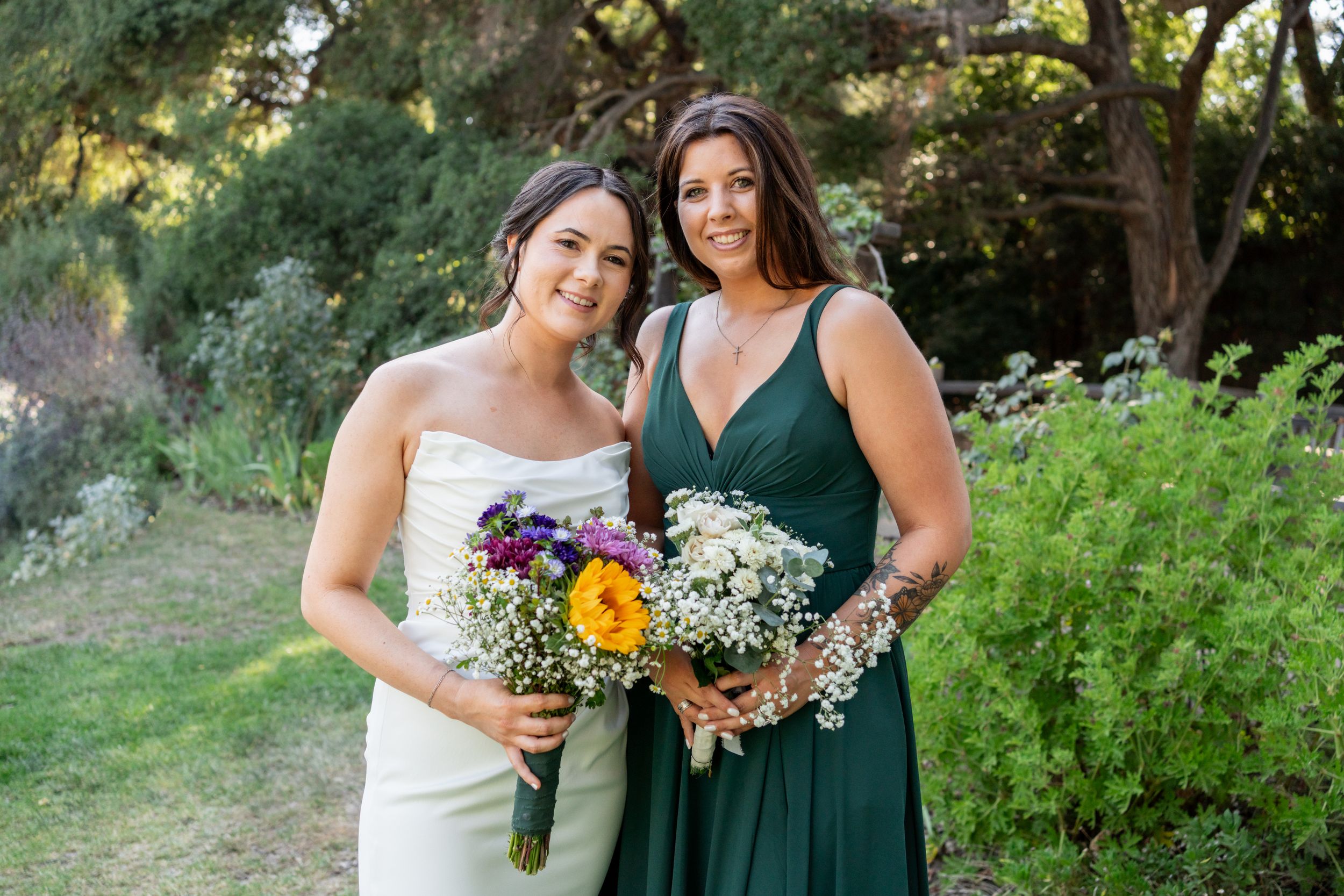 Bride and bridesmaid in green dress smile together holding colorful wildflower bouquets outdoors.