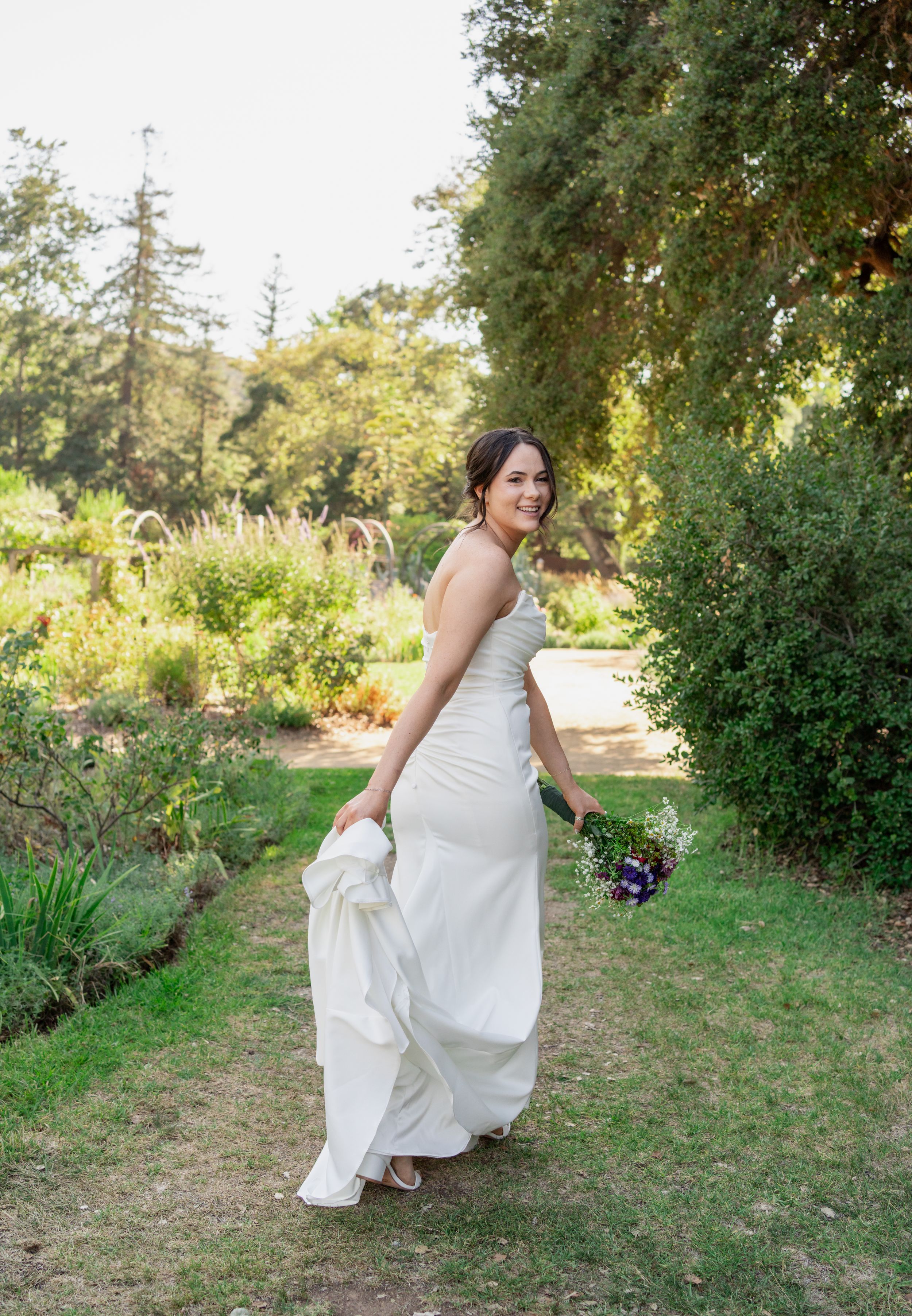 Bride in strapless ivory gown walks through lush garden holding small wildflower bouquet.