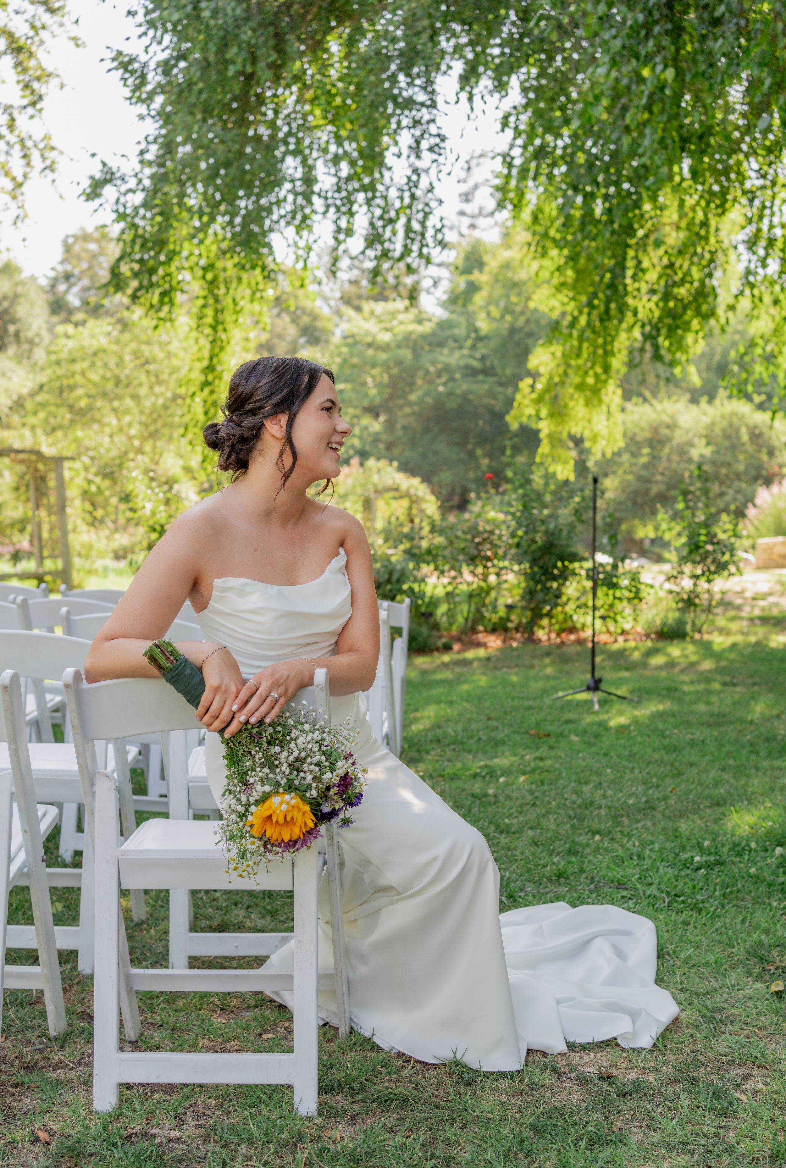 Smiling bride in strapless white gown sits on white chair holding colorful bouquet at outdoor garden wedding ceremony.