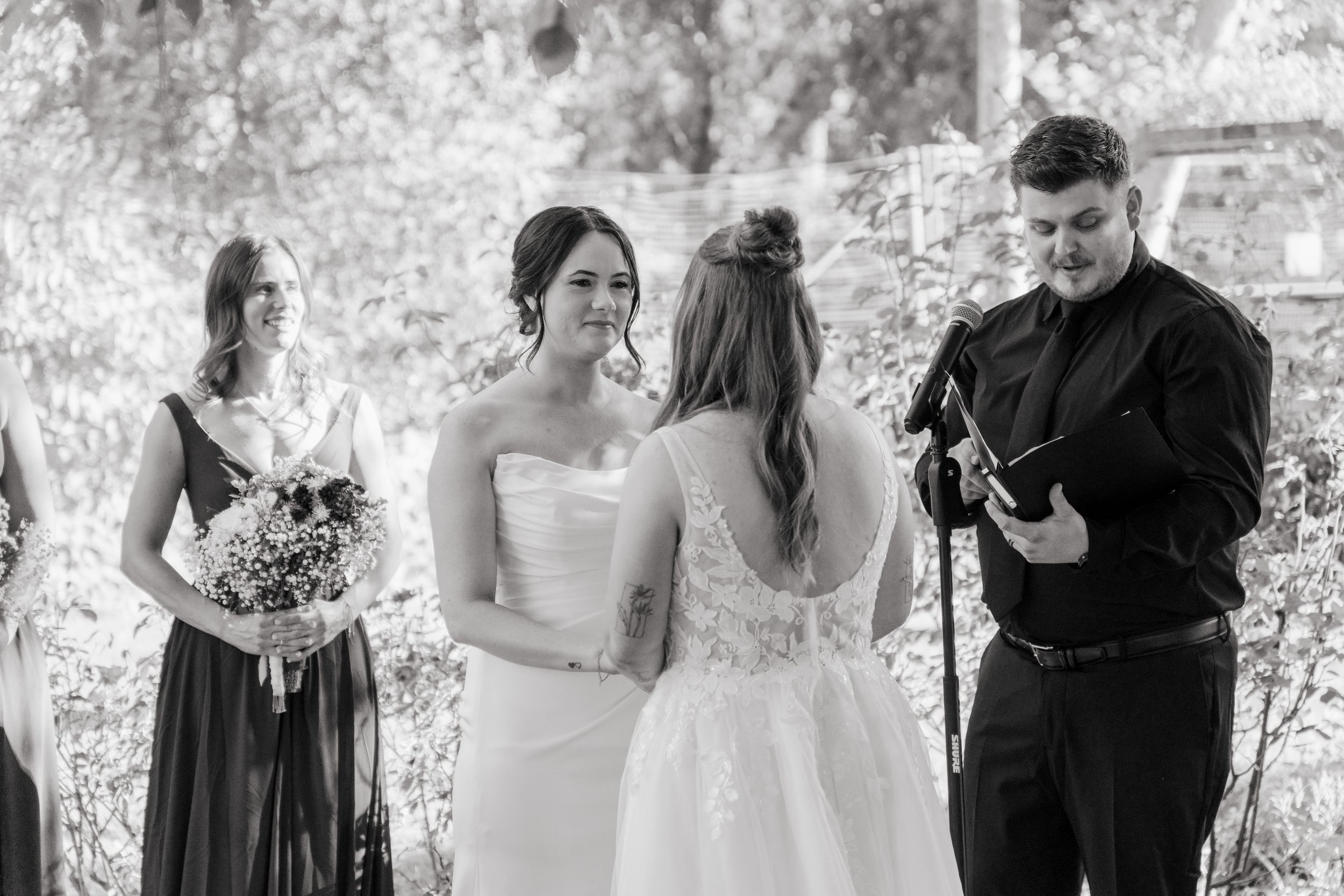 Two brides exchange vows at outdoor wedding ceremony in black and white photo with officiant and bridesmaid.