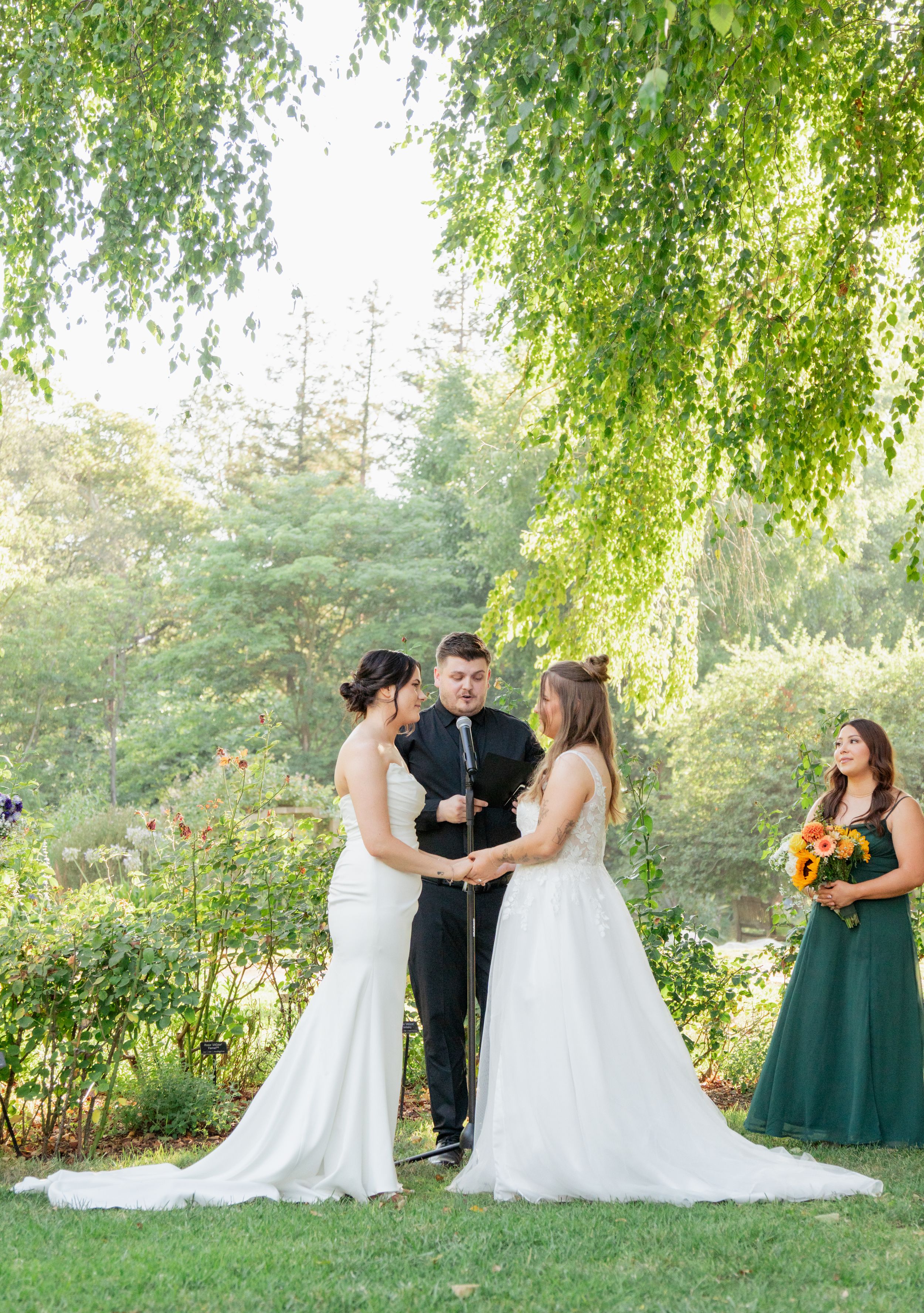 Two brides hold hands facing each other during outdoor garden wedding ceremony with officiant and bridesmaids.