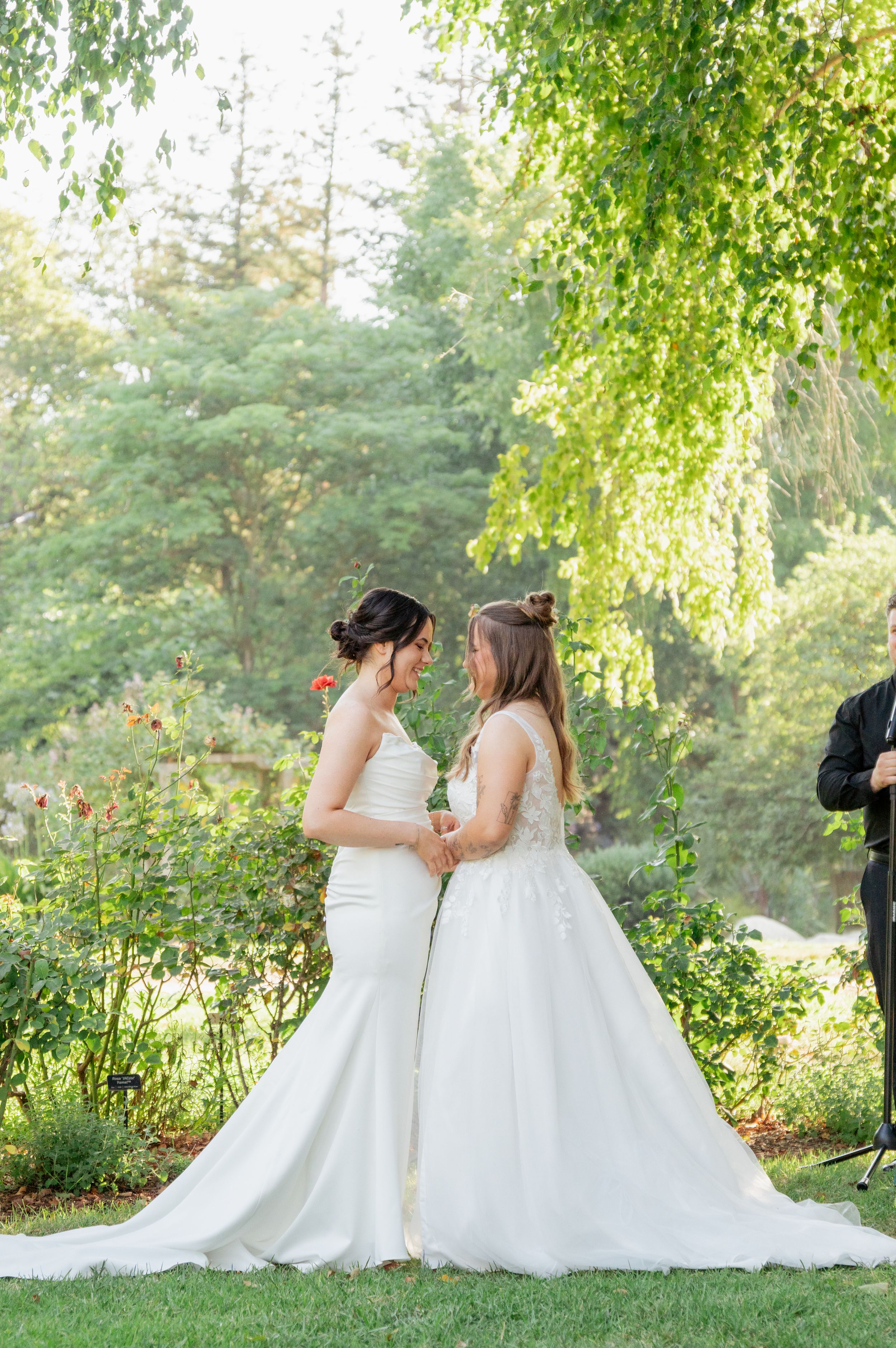 Two brides in white gowns hold hands and share an intimate moment under weeping willow tree at garden wedding.