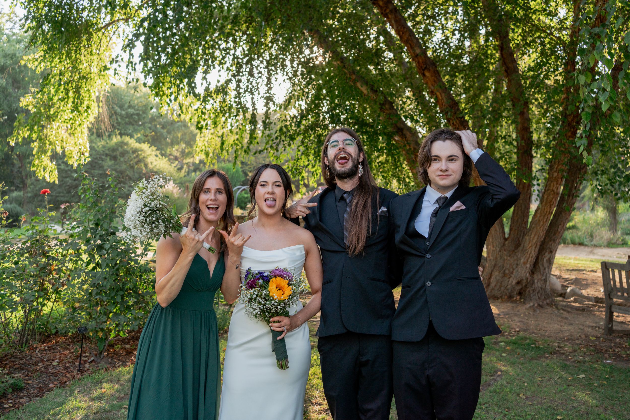 Bride and three wedding guests pose playfully together in lush green garden setting after outdoor ceremony.