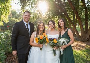 Two brides pose with family members holding colorful bouquets in golden sunlight at outdoor garden wedding.