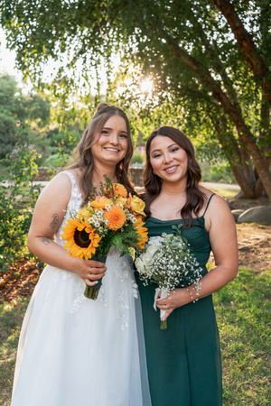 Bride in white gown and bridesmaid in green dress smile holding bouquets of sunflowers in garden at sunset.