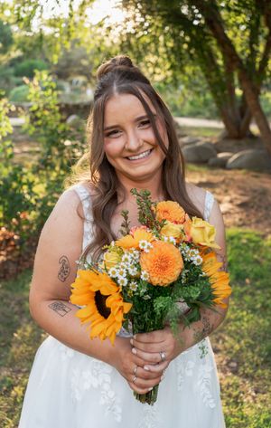 Smiling bride with half-up hairstyle holds vibrant sunflower and orange rose bouquet at outdoor garden wedding.