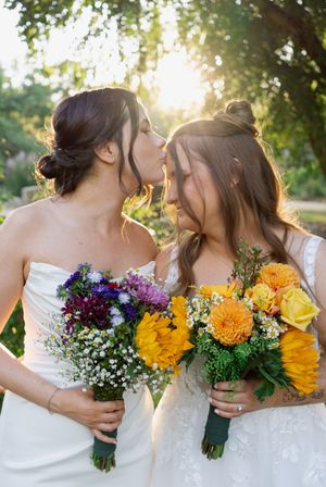Two brides share a kiss holding colorful bouquets with sunflowers and purple flowers in golden sunset light.