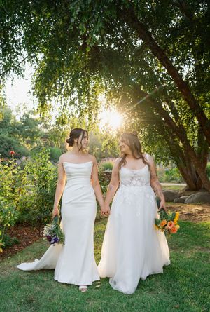 Two brides walk hand in hand in white wedding gowns through lush garden with golden sunset light behind them.