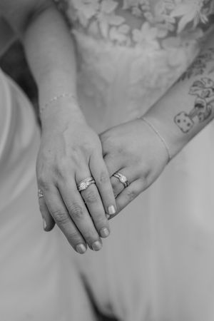 Close-up black and white photo of two brides holding hands showing wedding rings and bracelets in wedding dresses.