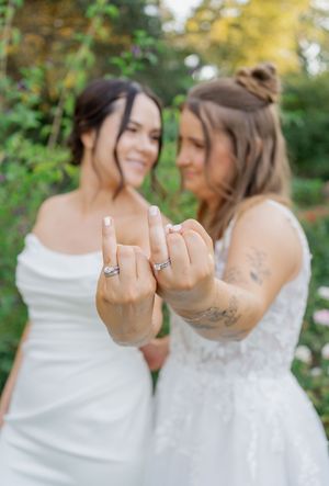 Two brides in white wedding dresses smiling and showing off their wedding rings in a garden setting.