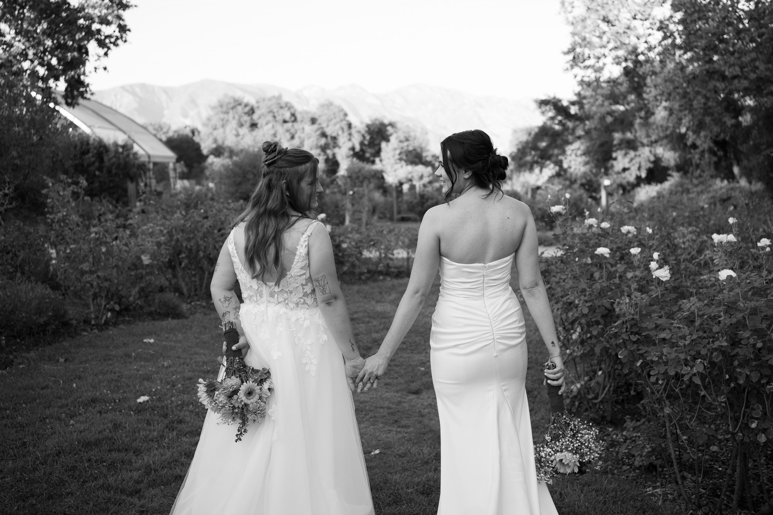 Black and white photo of two brides in white wedding dresses holding hands walking through a garden.