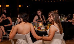 Two brides in white dresses sitting together at a candlelit wedding reception dinner table.