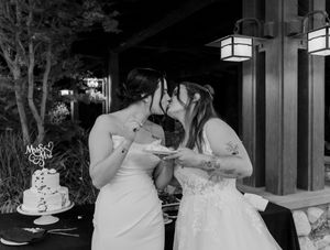 Black and white photo of two brides kissing while cutting their wedding cake at reception.