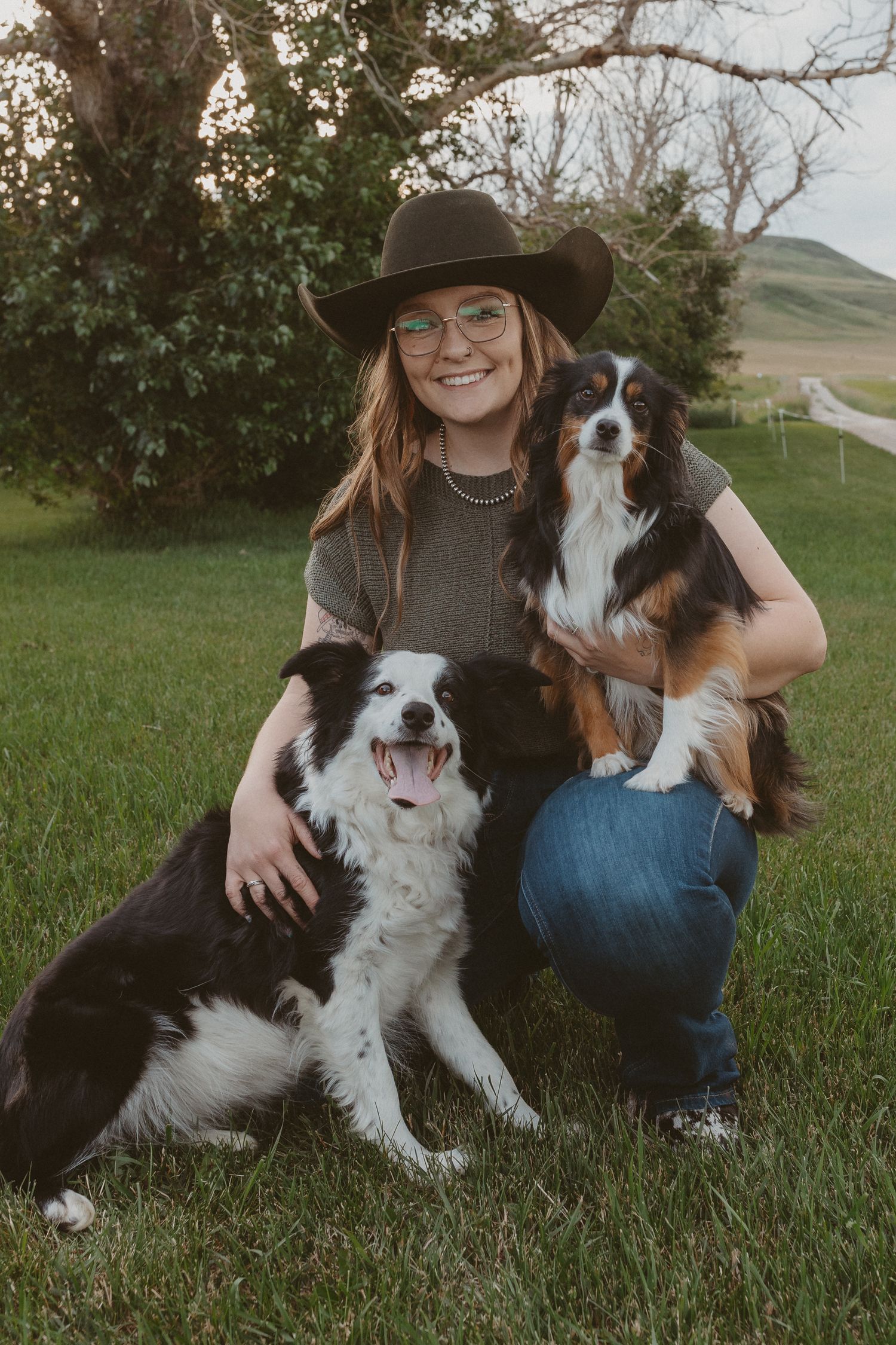 Someone in western wear poses with two happy border collies in a grassy yard.