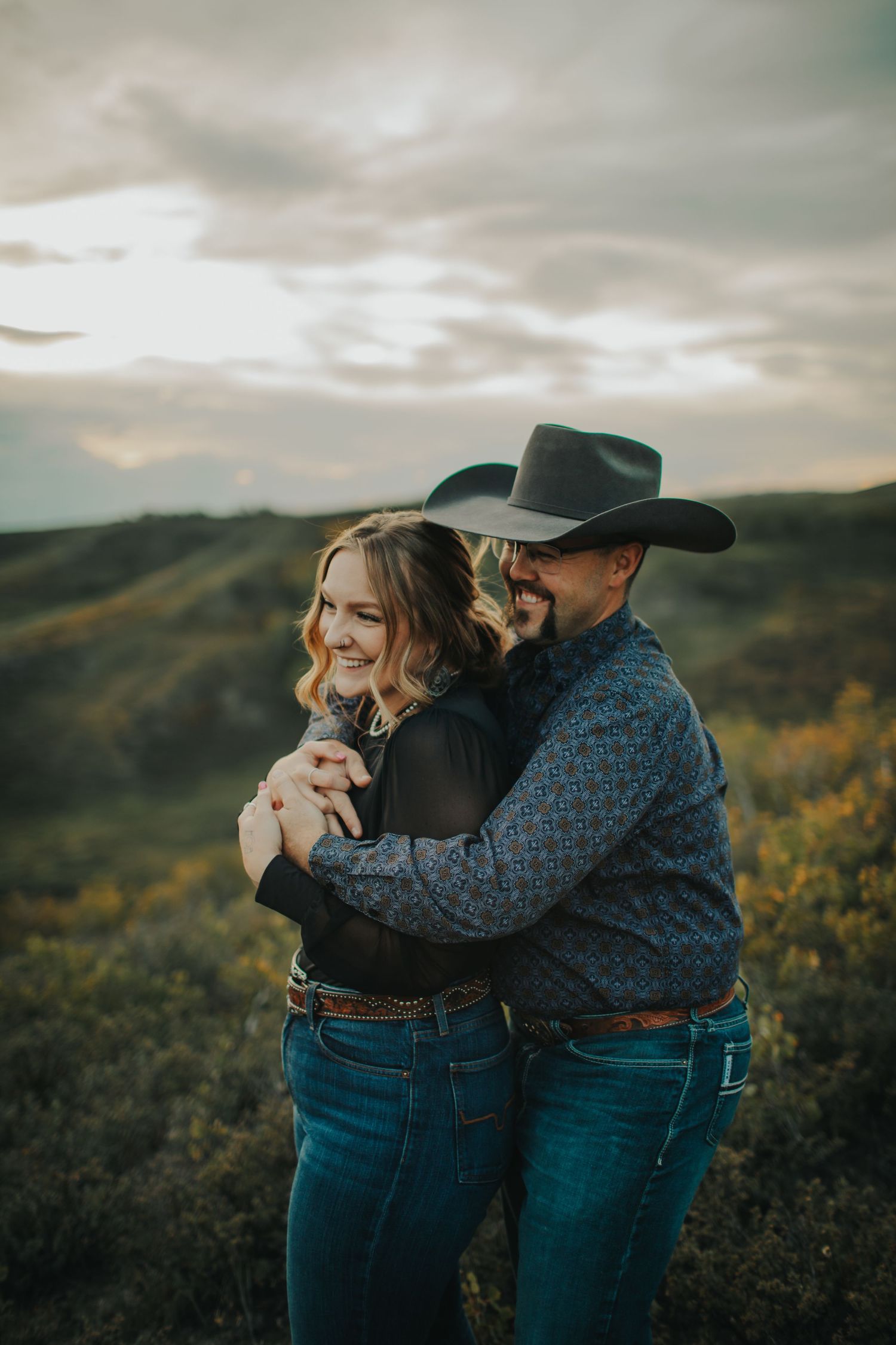 A couple embraces while wearing western attire against a sunset mountain backdrop.