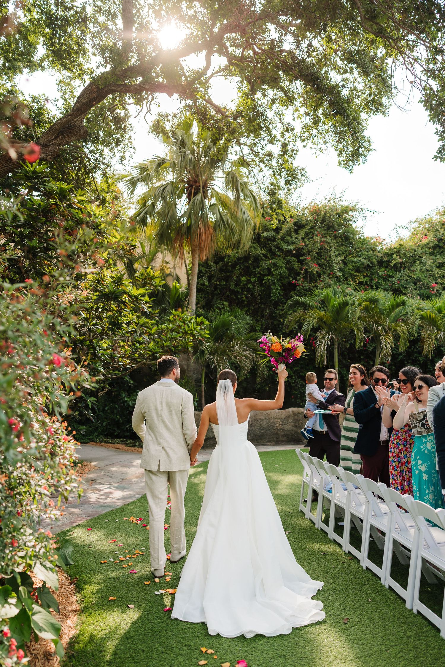 Bride and groom walk down outdoor aisle under lush trees, bride raising colorful bouquet, guests seated on white chairs.