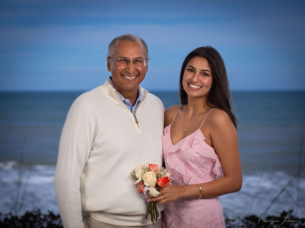 A couple poses at sunset on the beach with ocean waves in background, one holding a small bouquet of peach roses.