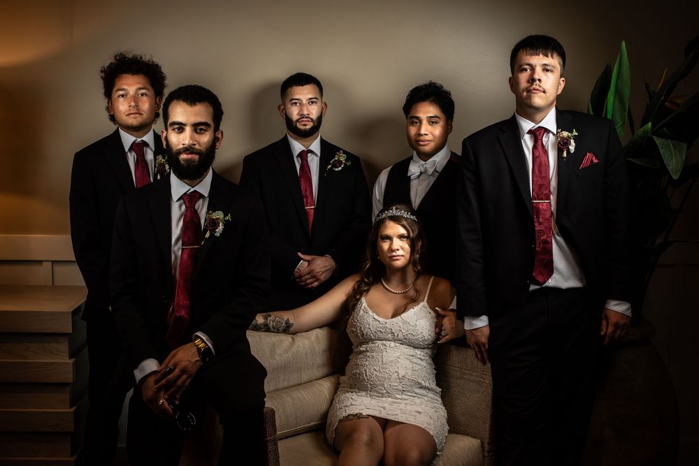 A moody wedding party portrait features a bride seated on a couch surrounded by groomsmen in dark suits and red ties.