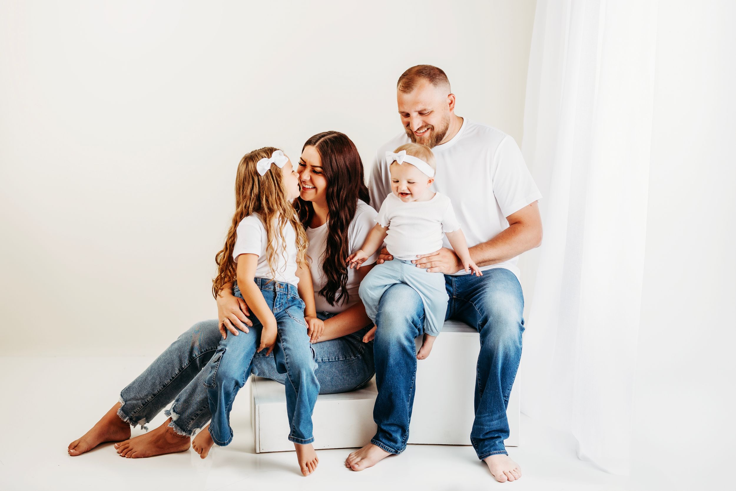 A family in casual denim outfits sits together on a white floor in a bright studio setting.