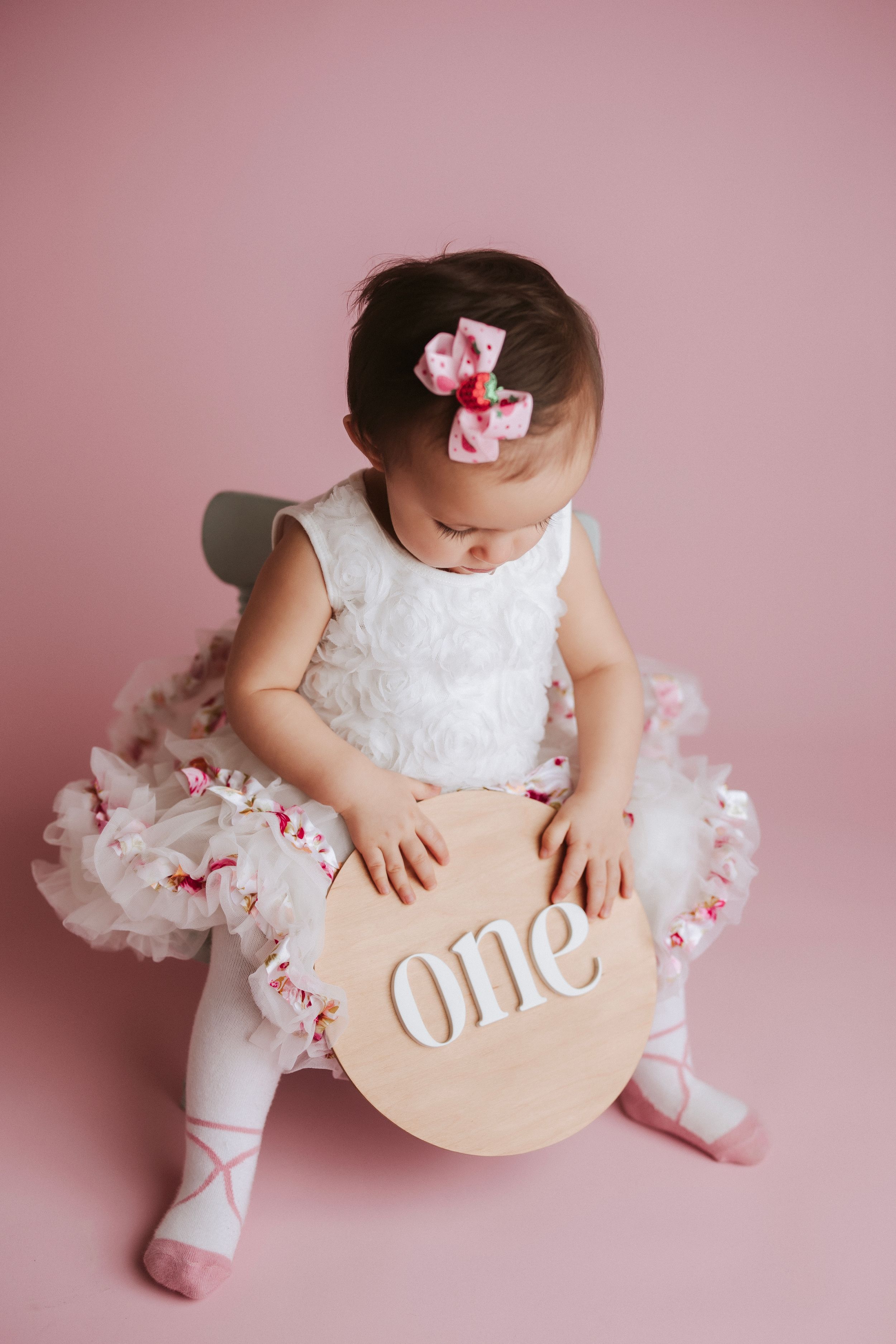 Baby in white lace dress and pink bow sits with wooden 'one' sign against pink backdrop in milestone photoshoot.