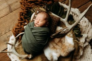Newborn baby wrapped in olive green swaddle lying on fur blanket with antlers in rustic setting.