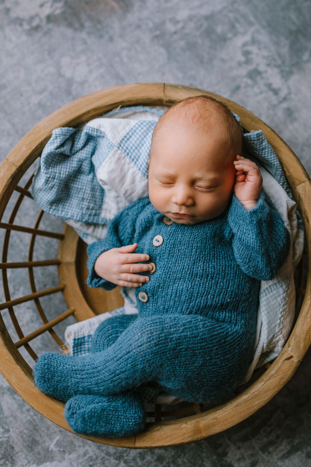 A sleeping newborn baby wearing a blue knit sweater lies peacefully in a round wooden basket.
