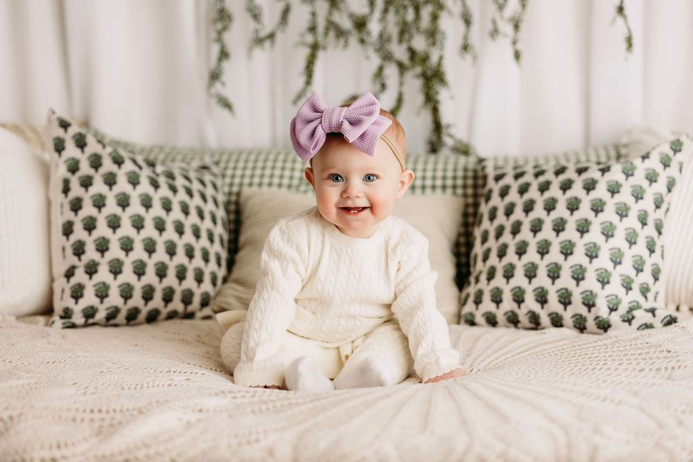 Baby in white outfit with purple bow headband sits on bed with patterned throw pillows.
