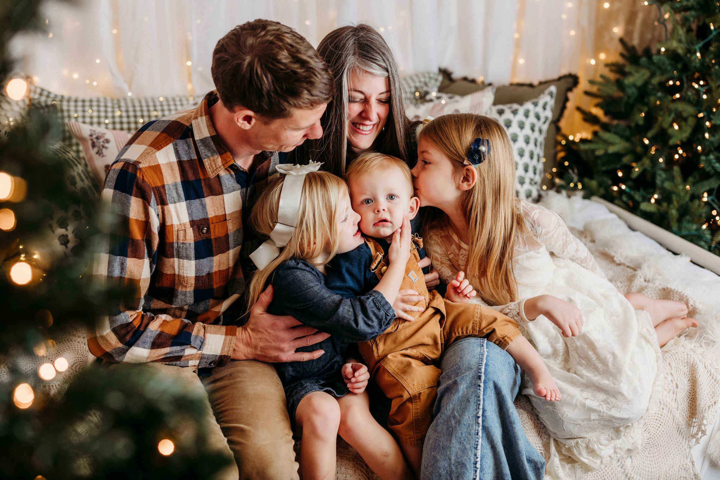 A family shares a joyful moment together on a couch near a Christmas tree with twinkling lights in the background.