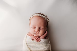 A sleeping newborn baby wrapped in white fabric with a delicate headband lies on a soft white backdrop.