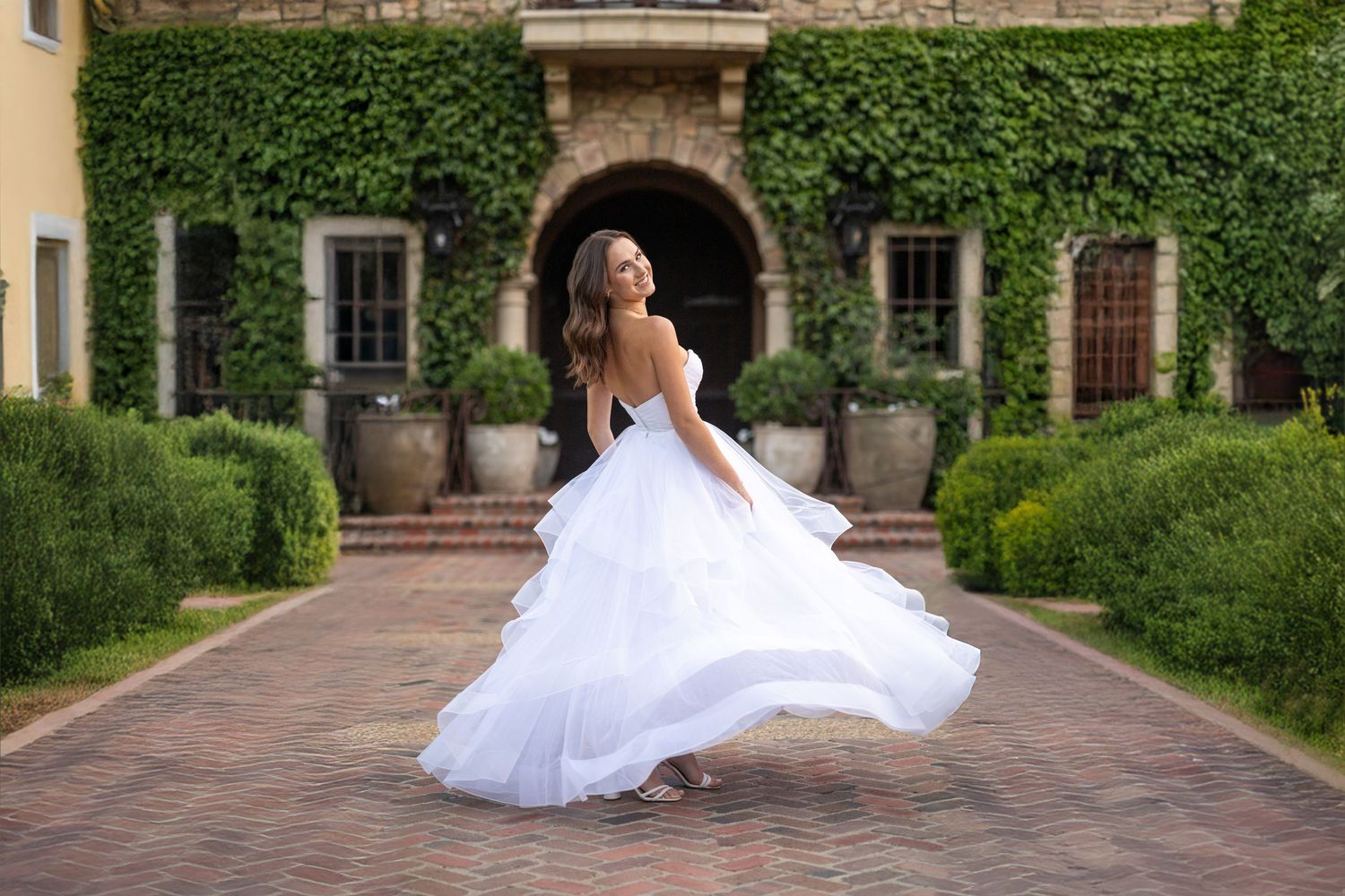 A high school senior in a white wedding gown and brown cowboy boots sits on stone steps near a colonial style building.