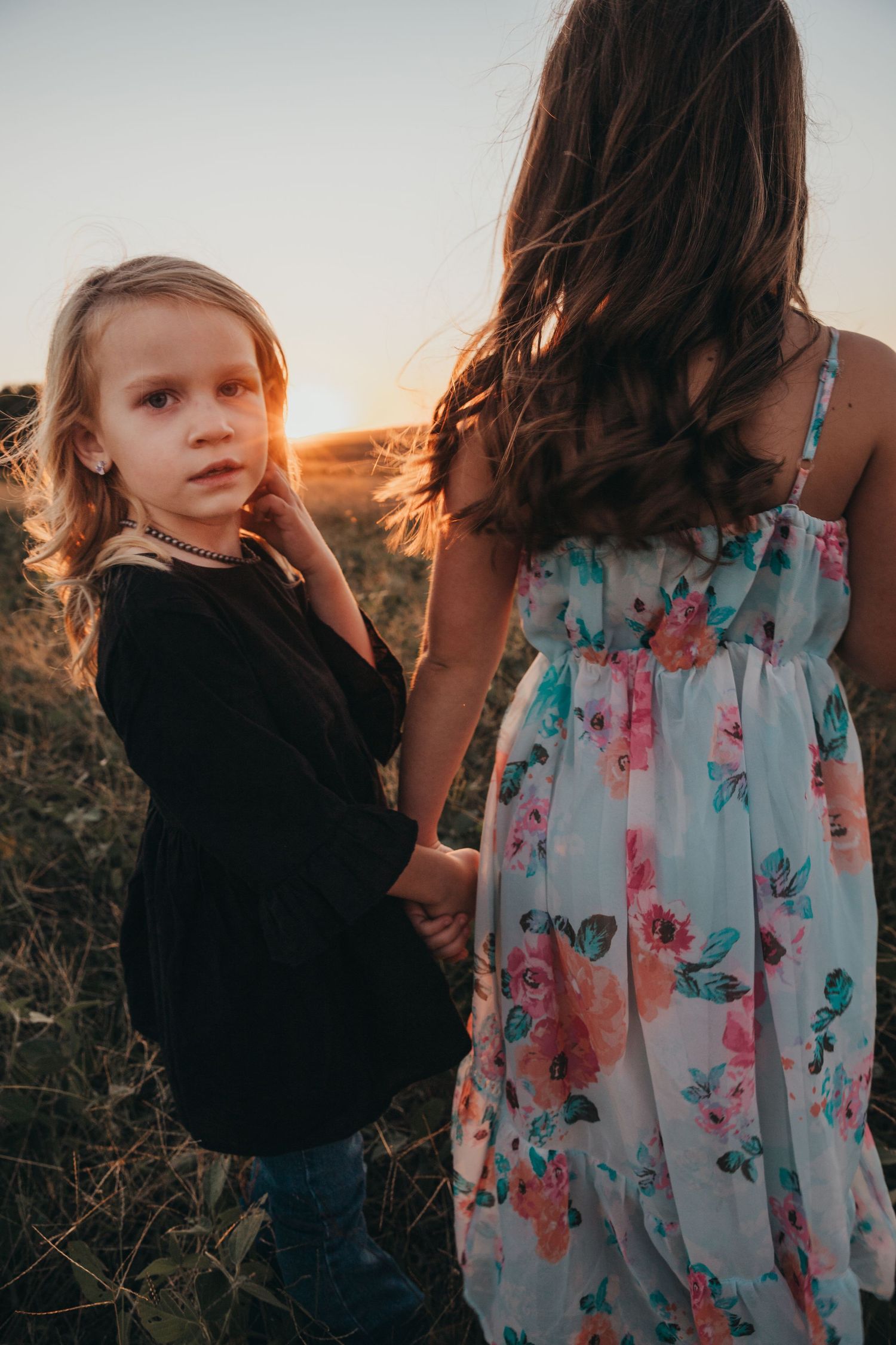 Child in black dress holding hands with person in floral dress during sunset.