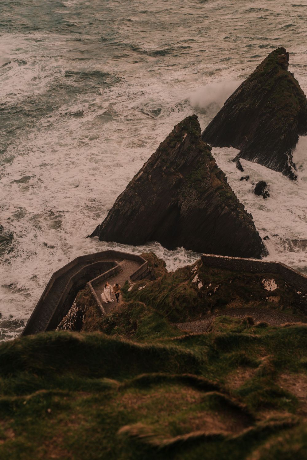 Dramatic aerial view of coastal cliffs with winding road and crashing waves below.