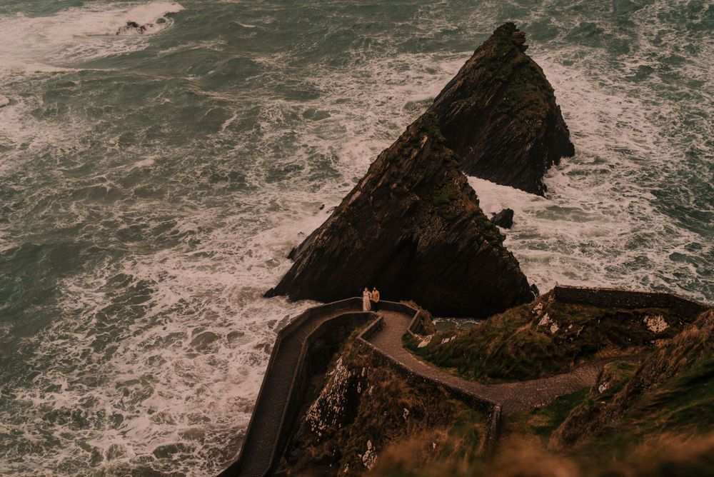 Aerial view of a dramatic coastal cliff with winding road path along rocky shoreline in moody lighting.