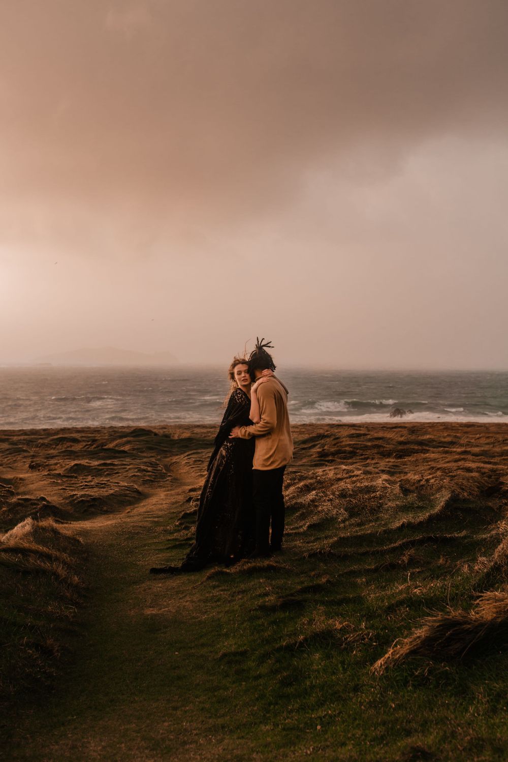 Couple stands together on windswept coastal grassland overlooking stormy ocean.