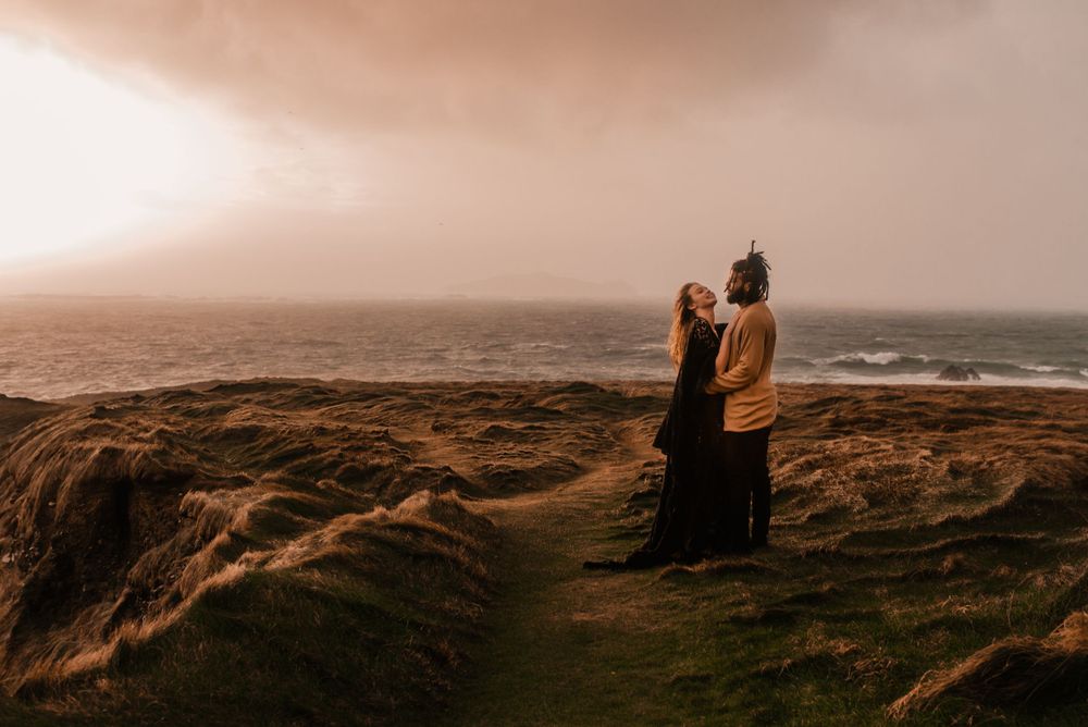 Romantic couple stands on windswept coastal cliffs during sunset in warm tones.