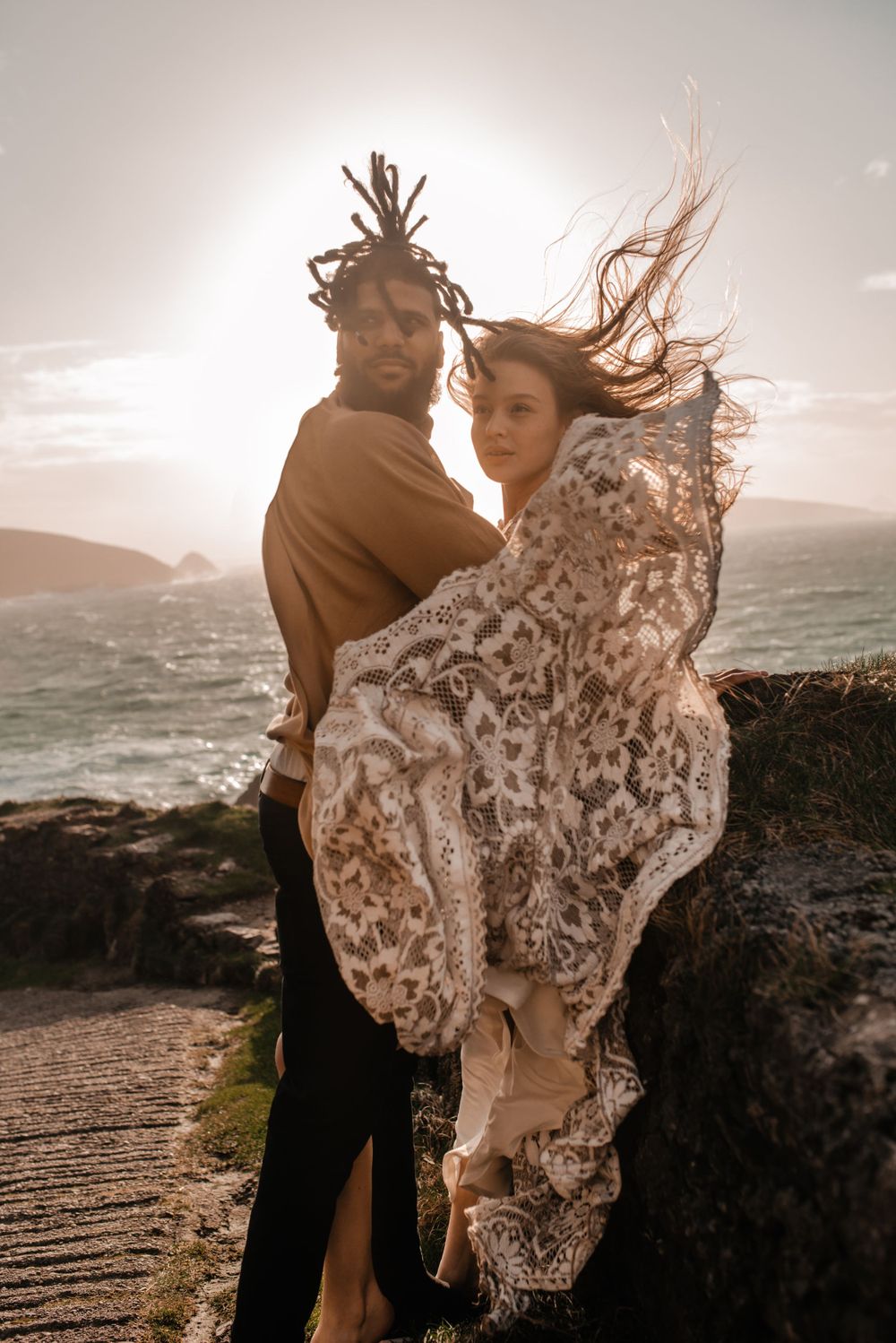 Couple in matching outfits share an intimate moment on a coastal overlook with white dress flowing.