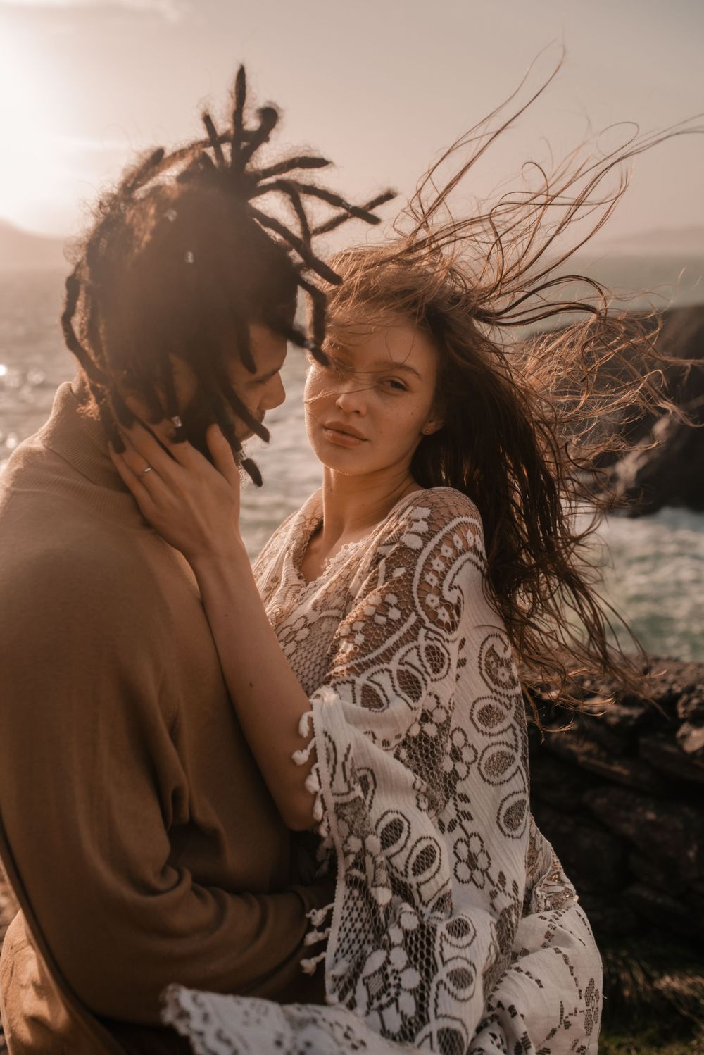A romantic couple embraces on a cliff overlooking the ocean at sunset with flowing hair in the wind.