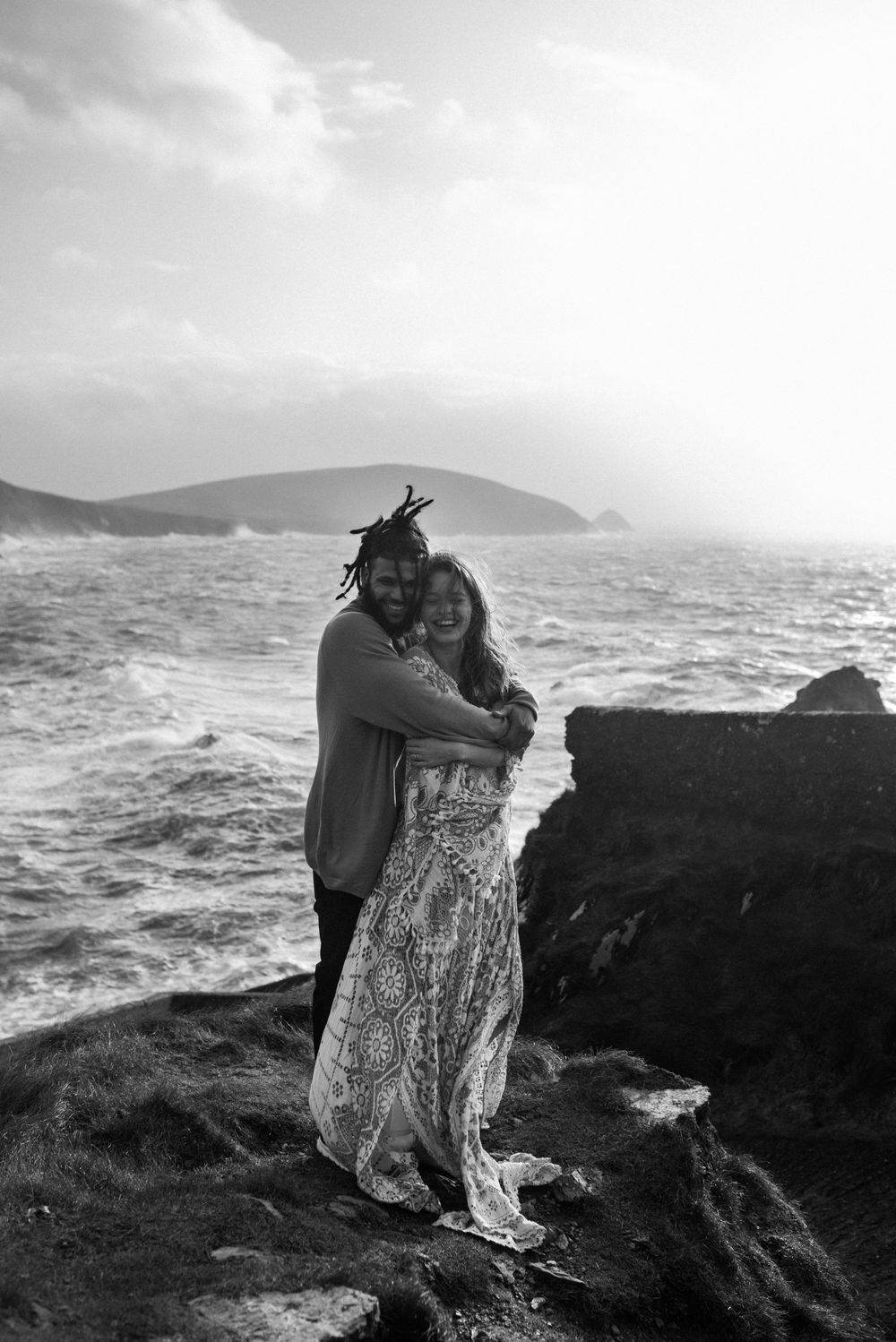 Couple embraces on rugged ocean cliffs during stormy weather in black and white photography.