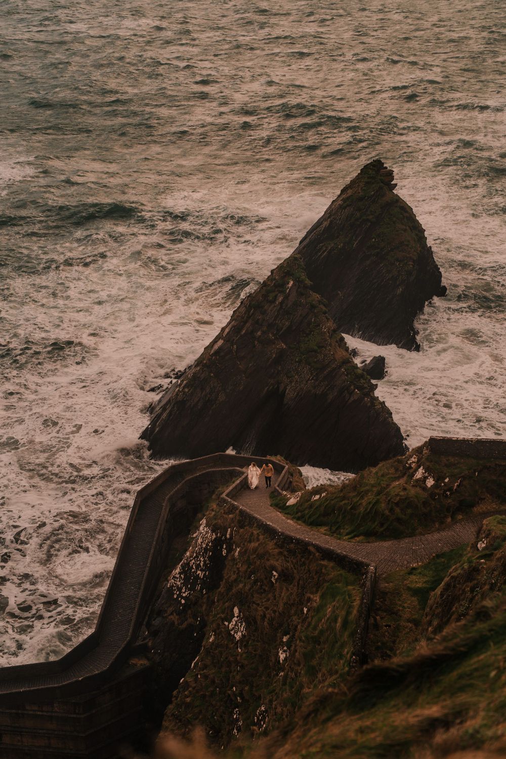 Dramatic coastal landscape with winding cliffside road and jagged rock formations jutting into churning ocean.